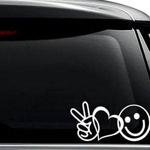 May include: White vinyl decal sticker with a peace sign, heart, and smiley face. The sticker is on a black car window.