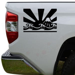 May include: Black vinyl decal featuring a stylized mountain, sun rays, and ocean waves design. The decal is applied to the side of a white vehicle. The design is a silhouette, with the mountain and waves at the bottom and the sun's rays at the top.