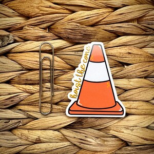 May include: A sticker featuring a cartoon traffic cone with the text "hoist the cone" in gold lettering. The cone is orange, white, and black. A silver paper clip is next to the sticker. The background is a woven, textured pattern.