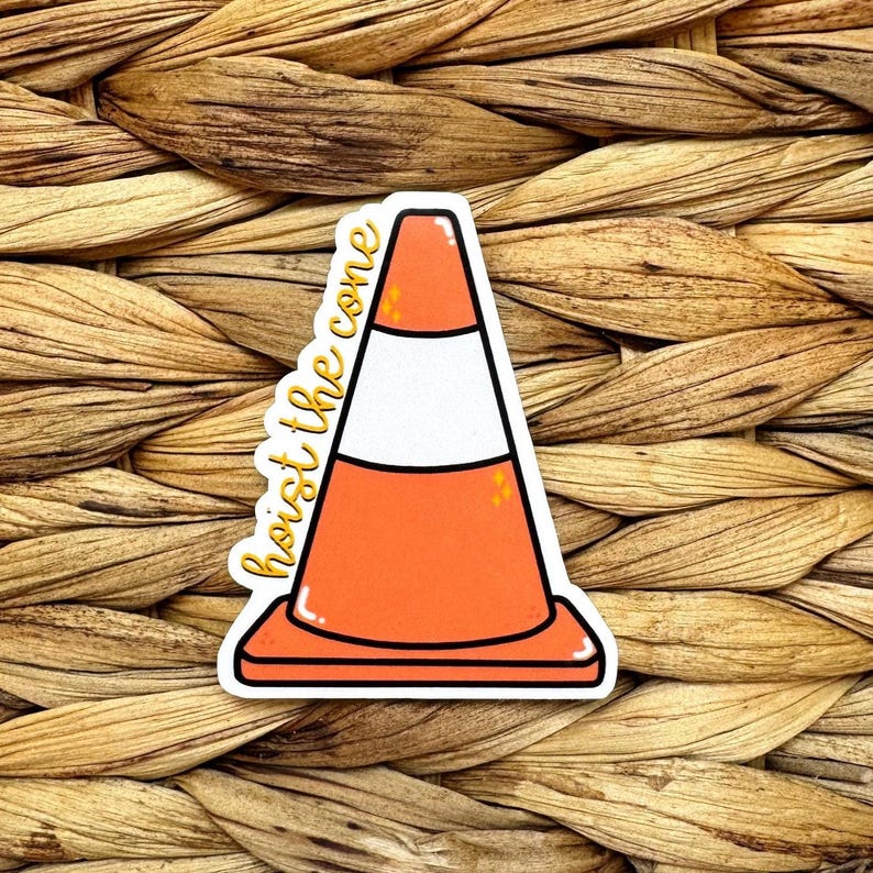 May include: A sticker featuring a cartoon traffic cone in orange, white, and black. The words "hoist the cone" are written in a cursive font on the left side of the cone. The sticker is on a woven background.
