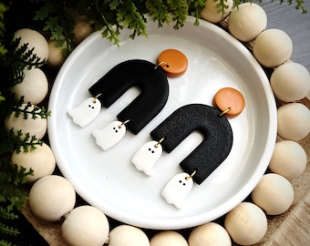 Könnte beinhalten: Ein Paar Halloween-Ohrringe. Jeder Ohrring hat ein schwarzes, strukturiertes, U-förmiges Design mit einem kleinen, weißen Geisteranhänger, der darunter hängt. Die Ohrringe haben eine runde, orangefarbene Oberseite.