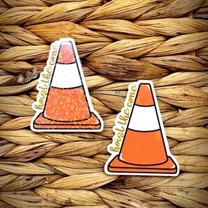 May include: Two stickers featuring orange and white traffic cones. One cone has glitter, the other is solid. The phrase "hoist the cone" is written in gold script on each sticker. The stickers are on a woven background.