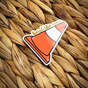 May include: A sticker featuring an orange and white traffic cone with the text "hoist the cone" in a handwritten style. The sticker has a black outline and is placed on a woven, textured surface.