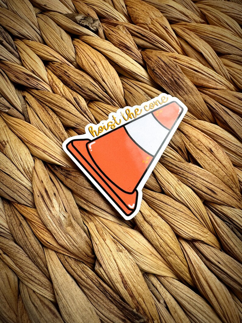 May include: A sticker featuring an orange and white traffic cone with the text "hoist the cone" in a handwritten style. The sticker has a black outline and is placed on a woven, textured surface.