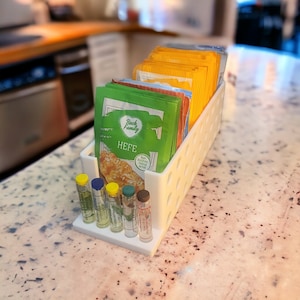 Könnte beinhalten: Weißer Plastik-Organizer mit kleinen Glasfläschchen und Hefe-Päckchen. Die Päckchen sind mit "Back Family Hefe" beschriftet und haben ein grün-gelb-weißes Design. Der Organizer hat ein Gitter aus Löchern zum Aufbewahren der Fläschchen und Päckchen.