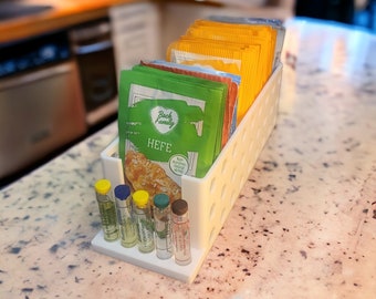 Op de afbeelding: Witte plastic organizer met kleine glazen flesjes en zakjes gist. De zakjes zijn gelabeld "Back Family Hefe" en hebben een groen, geel en wit ontwerp. De organizer heeft een raster van gaten voor het opbergen van de flesjes en zakjes.