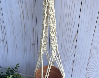 May include: A macrame plant hanger with a wooden ring at the top and a terracotta pot at the bottom. The hanger is made of natural cotton rope and has a fringe at the bottom.