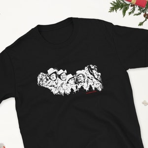 May include: Black t-shirt with a white graphic print of a group of people wearing cowboy hats. The graphic is a black and white illustration of a group of people with their arms around each other. The text "The Magnificent Seven" is printed in red below the graphic.