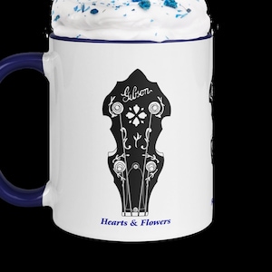 May include: White ceramic mug with a blue handle. The mug has a black and white illustration of a Gibson banjo headstock with the text "Hearts & Flowers" below it. The mug is filled with whipped cream and blue sprinkles and has a red and white striped straw.