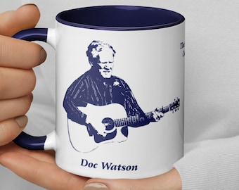 May include: White ceramic mug with a blue rim and handle. The mug features a black and white illustration of Doc Watson playing a guitar. The text "Doc Watson" is printed in blue below the illustration.