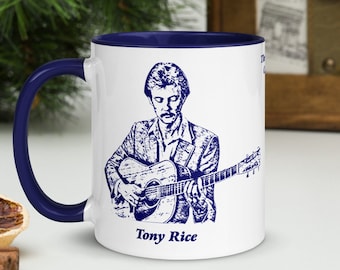 May include: White ceramic coffee mug with a blue rim and handle. The mug features a blue and white illustration of a man playing a guitar with the text "Tony Rice" below the image.