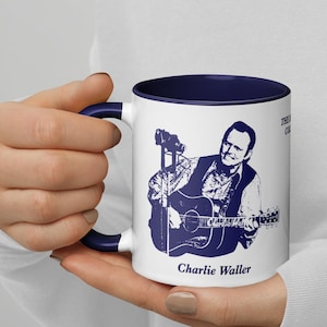 May include: White ceramic coffee mug with a blue rim and handle. The mug features a black and white illustration of Charlie Waller playing a guitar. The text "The Heart of Country Music" and "Charlie Waller" are printed on the mug.