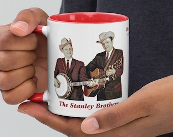 May include: White ceramic mug with a red rim. The mug features a photo of two men in cowboy hats playing musical instruments. The text "The Stanley Brothers" is printed in red at the bottom of the mug.