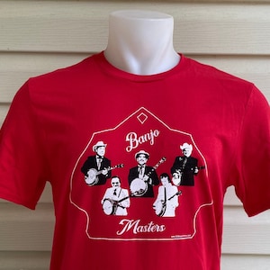 Op de afbeelding: Rood T-shirt met een witte afbeelding van een banjo-band in een huisvormig ontwerp. De tekst "Banjo Masters" staat onder de band. Het websiteadres "www.banjomasers.com" staat onder de tekst.