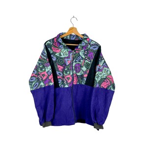 Może przedstawiać: Wintydowa kurtka polarowa z żywym geometrycznym wzorem w kolorze różowym, fioletowym, turkusowym i czarnym. Kurtka ma fioletową dolną część i czarne akcenty, z pełnym zapięciem na zamek i kołnierzem. Na drewnianym wieszaku.