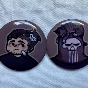 Pode incluir: Dois botões redondos com ilustrações de desenhos animados. Um botão apresenta uma pessoa com um gato na cabeça. O outro botão mostra uma caveira com um gato em cima. Ambos os botões têm um fundo castanho.