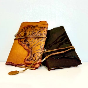 Puede incluir: Dos diarios encuadernados en cuero, uno marrón y otro tostado. El diario tostado presenta un mapa de la Tierra Media impreso en la portada. Ambos diarios están atados con correas de cuero.