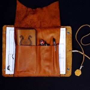 May include: A tan leather roll-up organizer featuring a flap, two front pockets, and a long cord tie. Inside are white paper sheets with Dungeons & Dragons text, a small tan notebook with a dragon emblem, a multi-colored die, and a silver pen.