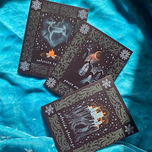 Peut inclure: Trois cartes de Noël marron foncé avec des illustrations hivernales. L'une représente un cerf et un feu de joie, une autre une église et un croissant de lune, et la troisième, des bougies allumées. Chaque carte a du texte et une bordure décorative avec des flocons de neige et de la verdure.