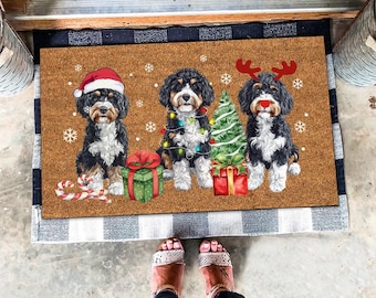 Peut inclure: Un paillasson en fibre de coco marron avec une bordure à carreaux blancs et noirs présente trois chiens doodle noirs et blancs portant des bonnets de Père Noël et des bois de renne. Les chiens sont debout devant un sapin de Noël avec des cadeaux et des lumières.