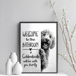 以下が含まれることがあります： 「WELCOME to the BATHROOM」の文字とゴールデンレトリバーが描かれた黒いフレームの壁掛けアート。 「a Goldendoodle will be with you shortly」の文字も含まれています。 白い花瓶と花の枝が前景にあります。