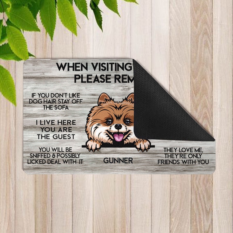 May include: A wooden welcome mat with a brown and white Pomeranian dog peeking over the top. The mat has the text "WHEN VISITING PLEASE REMEMBER" and "IF YOU DON'T LIKE DOG HAIR STAY OFF THE SOFA" and "I LIVE HERE YOU ARE THE GUEST" and "YOU WILL BE SNIFFED & POSSIBLY LICKED DEAL WITH IT" and "GUNNER" and "THEY LOVE ME, THEY'RE ONLY FRIENDS WITH YOU".