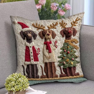 Könnte beinhalten: Besticktes Zierkissen mit drei Hunden in festlicher Kleidung. Einer trägt einen Weihnachtsmannhut und -mantel, ein anderer einen Schal, und der dritte einen Schal und hält einen Weihnachtsbaum. Das Kissen ist beige mit einem grünen Rand.