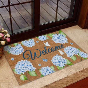 Peut inclure: Un paillasson rectangulaire avec un fond marron et un motif floral avec des hortensias bleus et des papillons. Le mot "Welcome" est écrit en bleu sur le tapis.