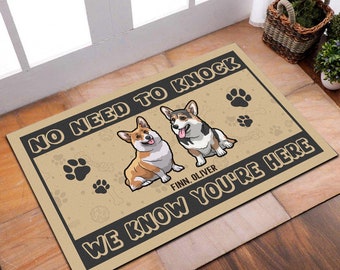 May include: A rectangular doormat with the text "NO NEED TO KNOCK WE KNOW YOU'RE HERE" in black. Two cartoon corgi dogs are featured, along with paw prints. The mat is tan with a black border.