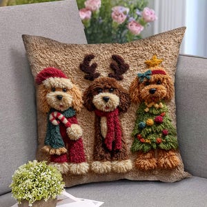 May include: A decorative square pillow featuring three dog figures in festive holiday attire. One dog wears a Santa hat and scarf, another has reindeer antlers and a scarf, and the third is dressed as a Christmas tree. The pillow has a textured, tan background.