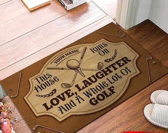 May include: A brown doormat with a woodgrain pattern and the text "This House Runs On Love, Laughter And A Whole Lot Of Golf". The doormat has a golf ball and golf clubs in the centre. The text "Your Name" is at the top of the doormat.