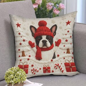 May include: A decorative Christmas-themed pillow featuring a French Bulldog wearing a red hat, scarf, and mittens. The pillow has a cream background with embroidered Christmas trees, gifts, and snowflakes. The dog is black and white.