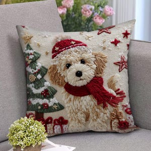 Könnte beinhalten: Besticktes Zierkissen mit einem hellbraunen Hund, der eine rote Mütze und einen Schal trägt. Ein Weihnachtsbaum und verpackte Geschenke sind ebenfalls auf dem Kissen gestickt. Rote Sterne sind auf dem cremefarbenen Hintergrund verstreut.