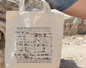 May include: A beige tote bag with a black and white print of a library card. The print includes the text "REPORT CHANGE OF ADDRESS PROMPTLY" and a table with dates and the words "DUE" and "RETURNED". The text at the bottom of the print reads "DO NOT LOSE THIS CARD, BOOKS DRAWN ON IT EITHER WITH OR WITHOUT YOUR CONSENT MUST BE AT YOUR RISK. LOSS OF CARD DOES NOT RELEASE YOU, EVEN IF REPORTED."