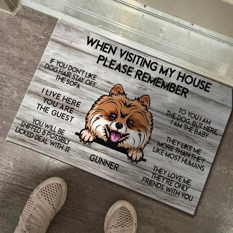 May include: A welcome mat with a woodgrain background and a cartoon image of a brown and white Pomeranian dog peeking over the top. The text on the mat reads "When visiting my house please remember..." and includes a list of rules for visitors, such as "To you I am the dog, but here, I am the baby." and "They love me, they're only friends with you."