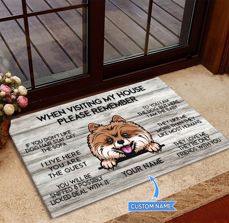 May include: A welcome mat with a brown and white dog peeking over the top. The mat has a woodgrain background and the text "When visiting my house please remember" and "To you I am the dog, but here I am the baby" and "They like me more than they like most humans" and "They love me, they're only friends with you".
