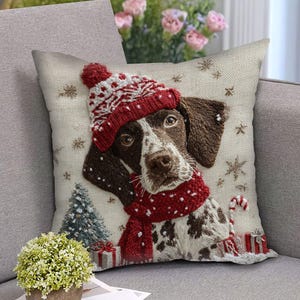Könnte beinhalten: Ein dekoratives quadratisches Kissen mit einem Hund, der eine rot-weiß gestrickte Mütze und einen Schal trägt. Der Hund hat braunes und weißes Fell, umgeben von Schneeflocken, einem Weihnachtsbaum und Geschenken. Das Kissen liegt auf einem grauen Sofa.