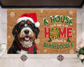 Peut inclure: Tapis de porte rectangulaire avec un chien Bernedoodle portant un bonnet de Père Noël et une écharpe rouge et verte. Le tapis affiche le texte "A HOUSE is not a HOME without a BERNEDOODLE" en vert et rouge, avec des flocons de neige.