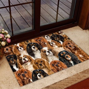 Op de afbeelding: Een deurmat met een patroon van veel Cavalier King Charles Spaniel honden in verschillende poses. De honden zijn bruin, zwart en wit.
