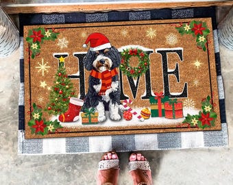 Peut inclure: Un paillasson en coco brun avec le mot "HOME" en lettres blanches. Un chien noir et blanc portant un bonnet de Père Noël rouge se tient devant le mot "HOME". Le paillasson est décoré d'éléments de Noël, notamment un sapin de Noël, des cadeaux et des cannes en sucre.