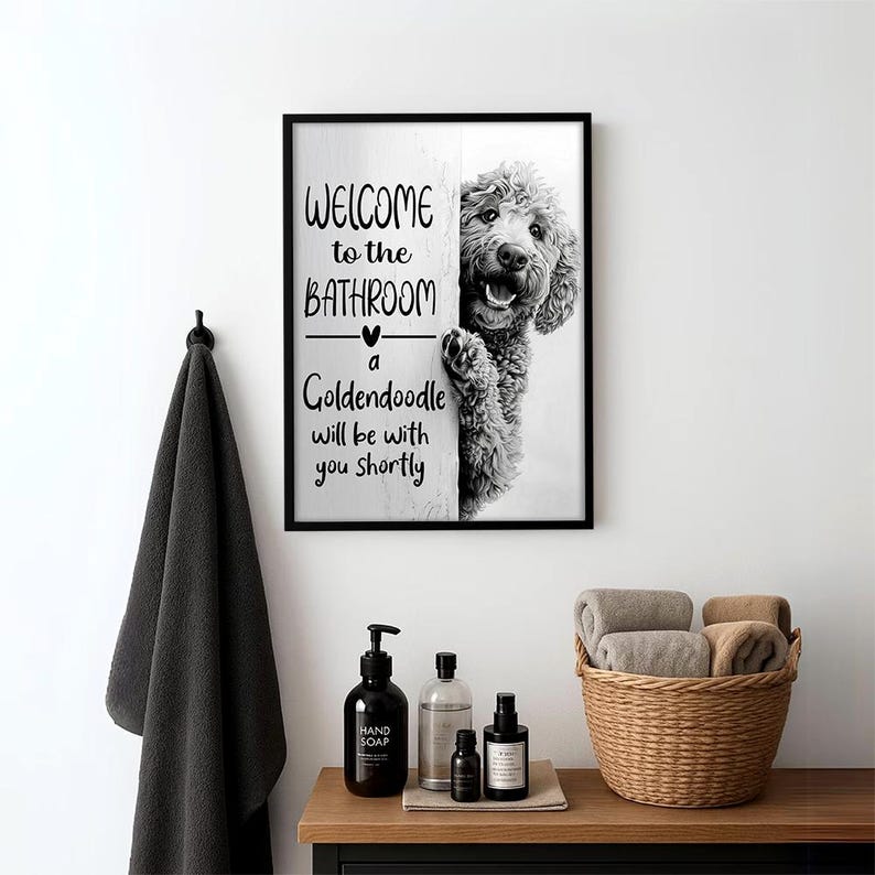 以下が含まれることがあります： 「WELCOME to the BATHROOM a Goldendoodle will be with you shortly」の文字とゴールデンレトリバーの犬が描かれた白黒のフレーム入りプリント。黒いタオルがフックに掛けられ、タオルのバスケットが木製の棚に置かれています。