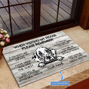 Op de afbeelding: Een houten deurmat met een Dalmatische hond die over de bovenkant kijkt. De deurmat heeft de tekst "When visiting my house please remember" en "To you I am the dog, but here I am the baby".