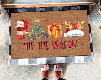 Può includere: Un tappetino per porta in fibra di cocco marrone con il testo "'Tis the Season" in lettere rosse. Il tappetino presenta un'illustrazione di cartone animato di una tazza di caffè con un cappello di Babbo Natale, un albero di Natale, un regalo avvolto e un cappello di Babbo Natale con le corna di renna.