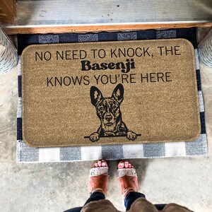 May include: A brown doormat with the text "No need to knock, the Basenji knows you're here" and a black and white illustration of a Basenji dog peeking over the top.