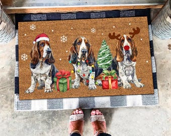 Op de afbeelding: Een bruine kokosvezel deurmat met drie basset hounds die kerstmanmutsen en rendiergewei dragen. De honden zijn omringd door kerstverlichting, cadeaus en een kerstboom. De deurmat heeft een geruite rand.