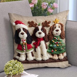 May include: A decorative square pillow featuring three dog figures in festive holiday attire. One dog wears a Santa hat and coat, another has reindeer antlers and a scarf, and the third is dressed as a Christmas tree. The pillow has a textured, beige background.