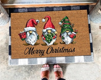 Può includere: Un tappetino per porta in fibra di cocco marrone con tre gnomi che indossano cappelli rossi e verdi. Gli gnomi tengono un albero di Natale, un regalo e una calza. Il testo "Merry Christmas" è stampato sul tappetino.