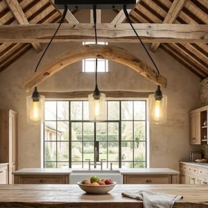 May include: Rustic kitchen lighting fixture with three pendant lights. The lights hang from a wooden beam and feature clear glass shades with Edison-style bulbs. The kitchen has a large window and wooden cabinets.