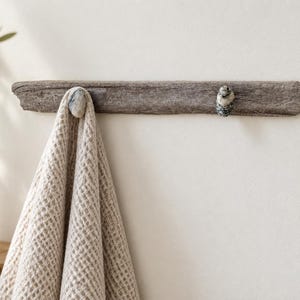 May include: A wall-mounted rack made from a weathered gray piece of driftwood featuring two smooth, rounded stone hooks. A beige, textured waffle-weave towel hangs from the left stone hook, displaying a natural, organic aesthetic.