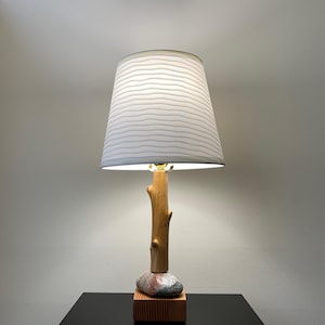 May include: A unique table lamp with a natural wood base and stem, topped with a white lampshade. The lamp features a stone and wooden block base. The lampshade has a horizontal striped pattern. The lamp is turned on, illuminating the room.